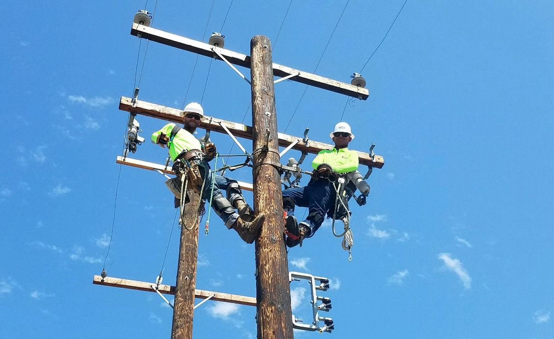 Two workers in safety gear are perched on a utility pole under a clear blue sky.