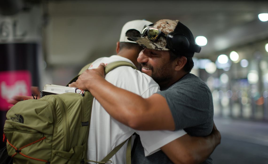 Two people embracing, one wearing a gray shirt and a cap with glasses on top, the other in white with a green backpack.