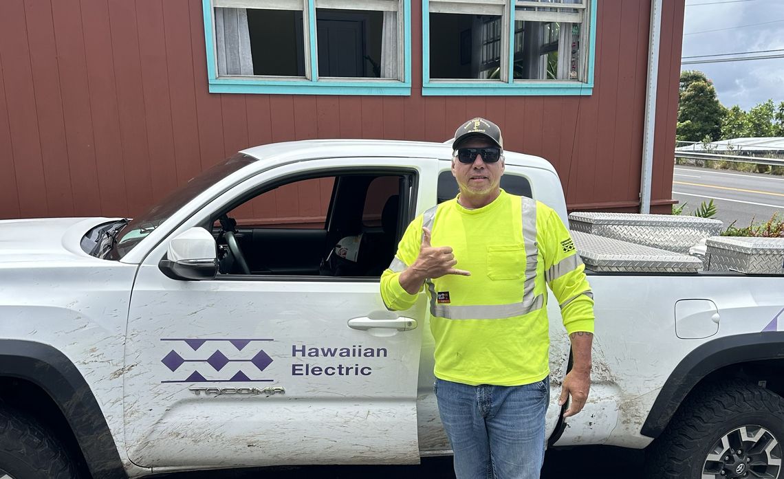 A person in a neon yellow shirt stands next to a Hawaiian Electric truck making a shaka sign.