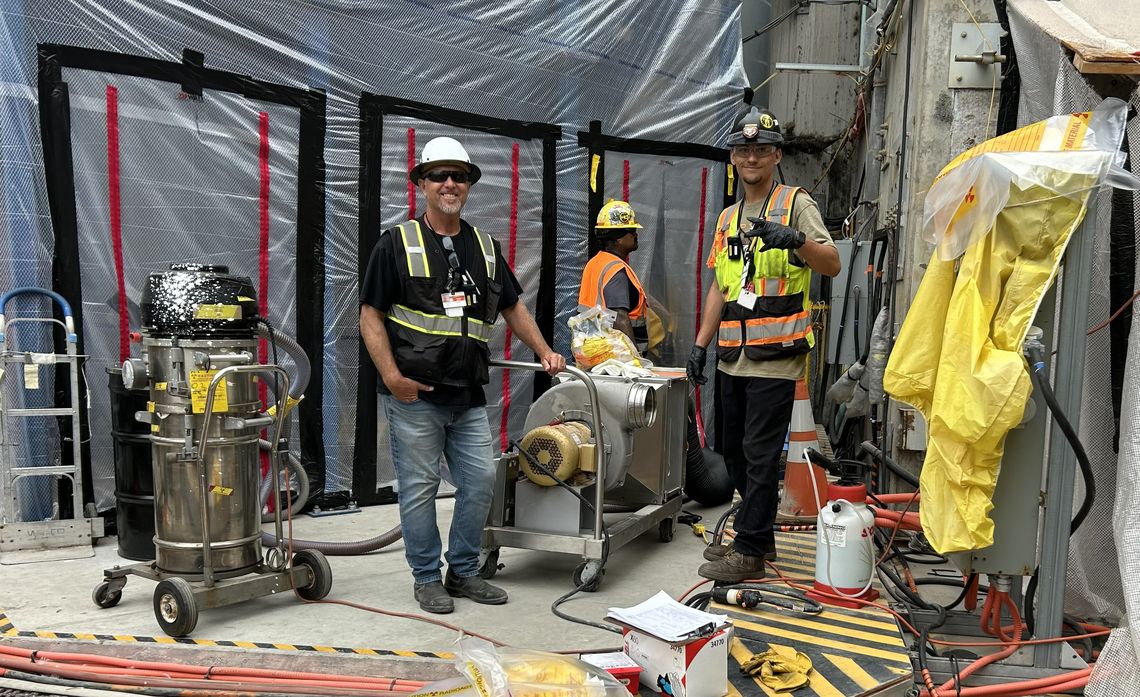 Three workers in safety gear stand inside an industrial site surrounded by machinery, caution tape, and protective tarps.