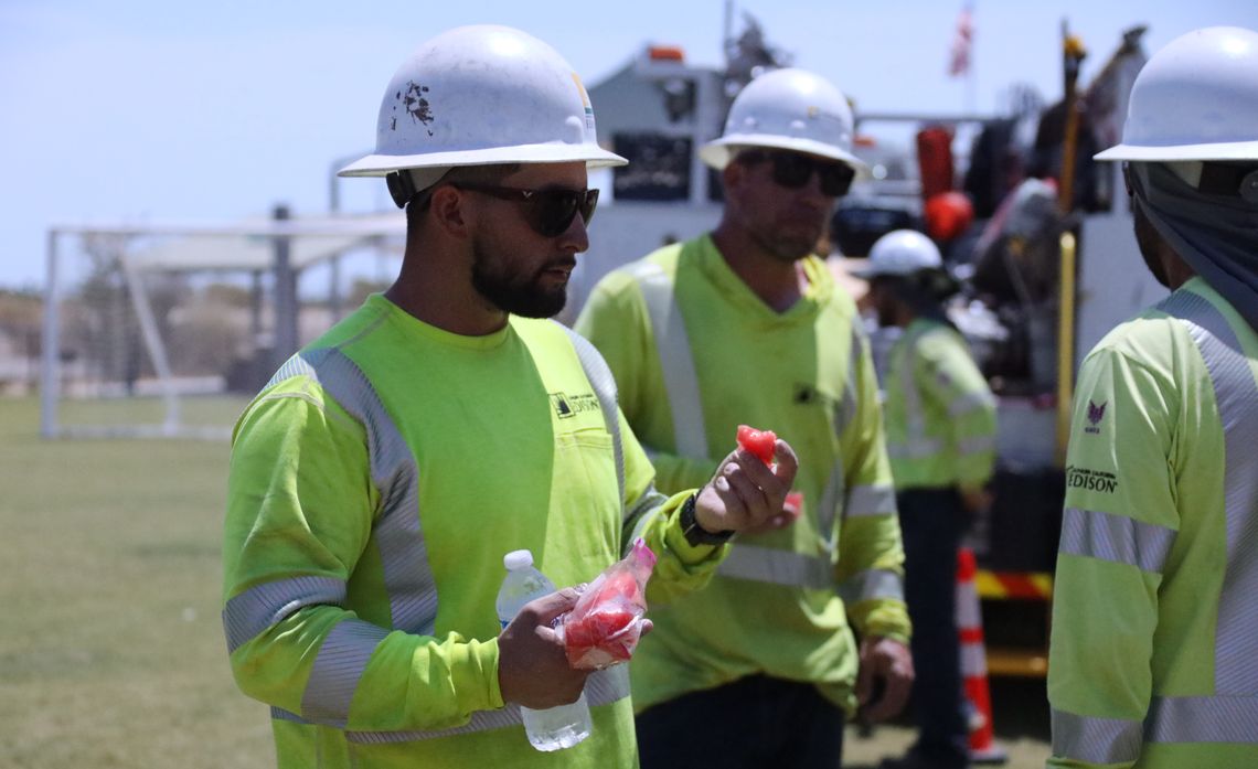 Utility workers in yellow safety gear and helmets eating watermelon outdoors.