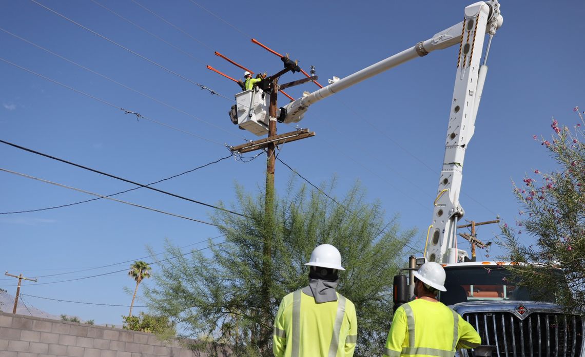 Utility workers in safety gear observe a colleague in a bucket lift working on high power lines against a clear blue sky.