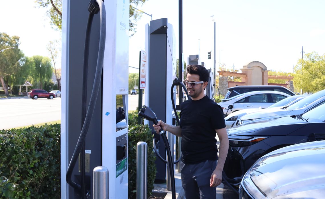 A man in sunglasses holds an electric vehicle charging connector beside a row of fast-charging stations and parked cars.