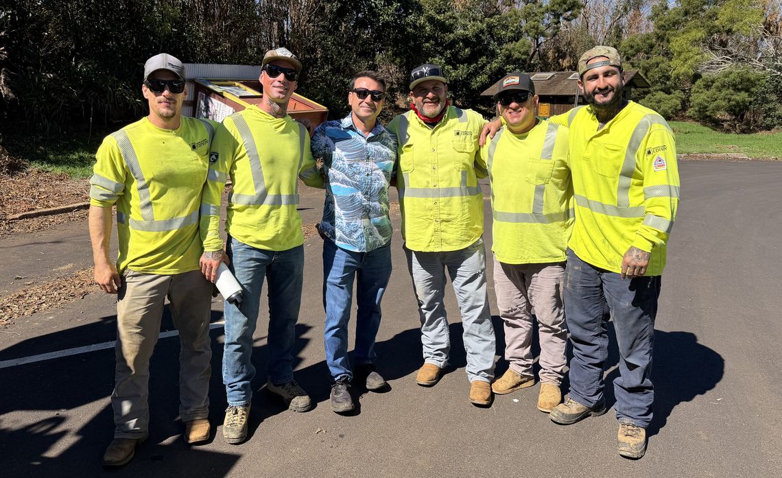 Six men, five wearing bright yellow high-visibility work uniforms, pose together outdoors on a sunny paved road.