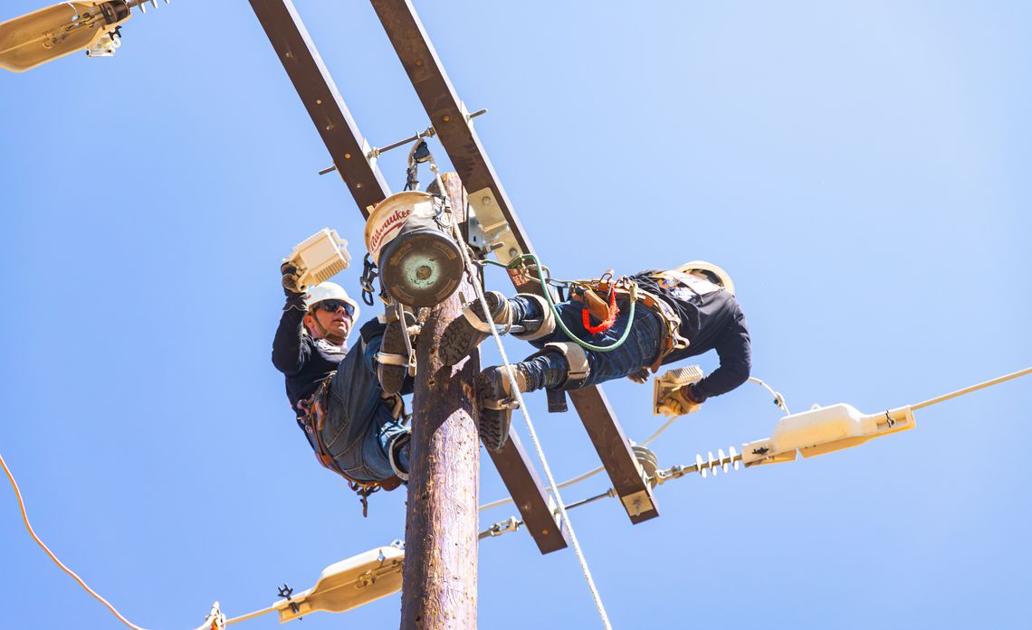 Two utility workers in safety gear climb a wooden power pole, repairing equipment and electrical lines against a clear blue sky.
