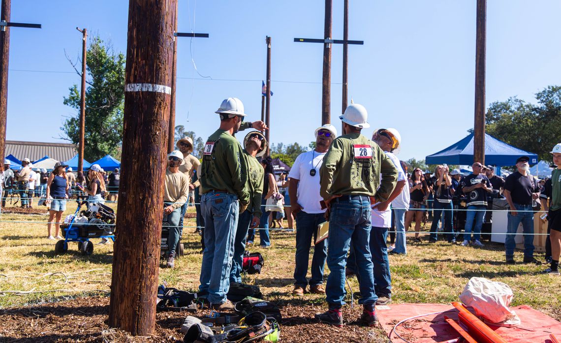 Utility workers in hard hats gather around wooden poles at an outdoor pole-climbing competition, with spectators and tents behind them.