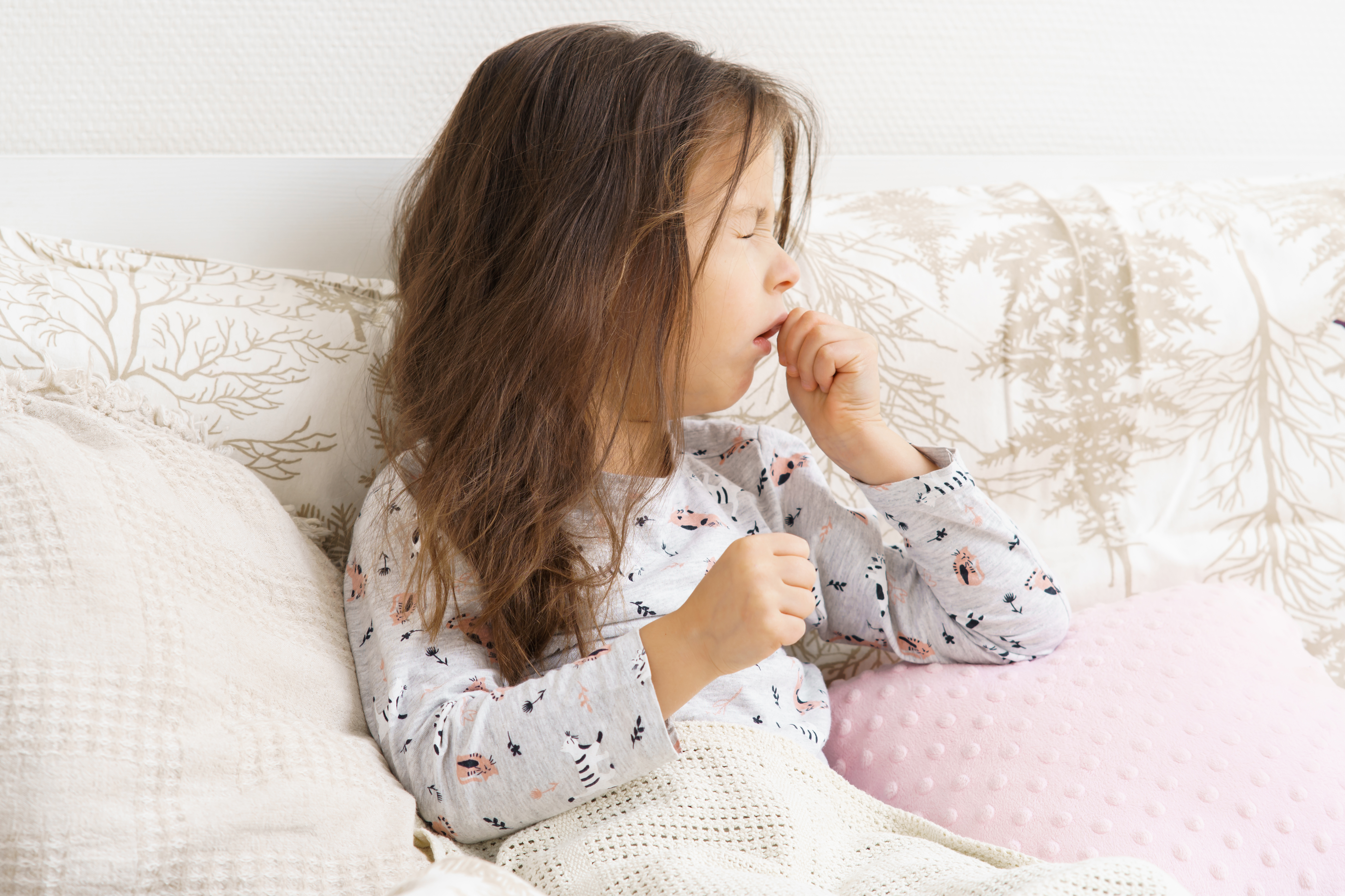 A young girl coughs while sitting in bed