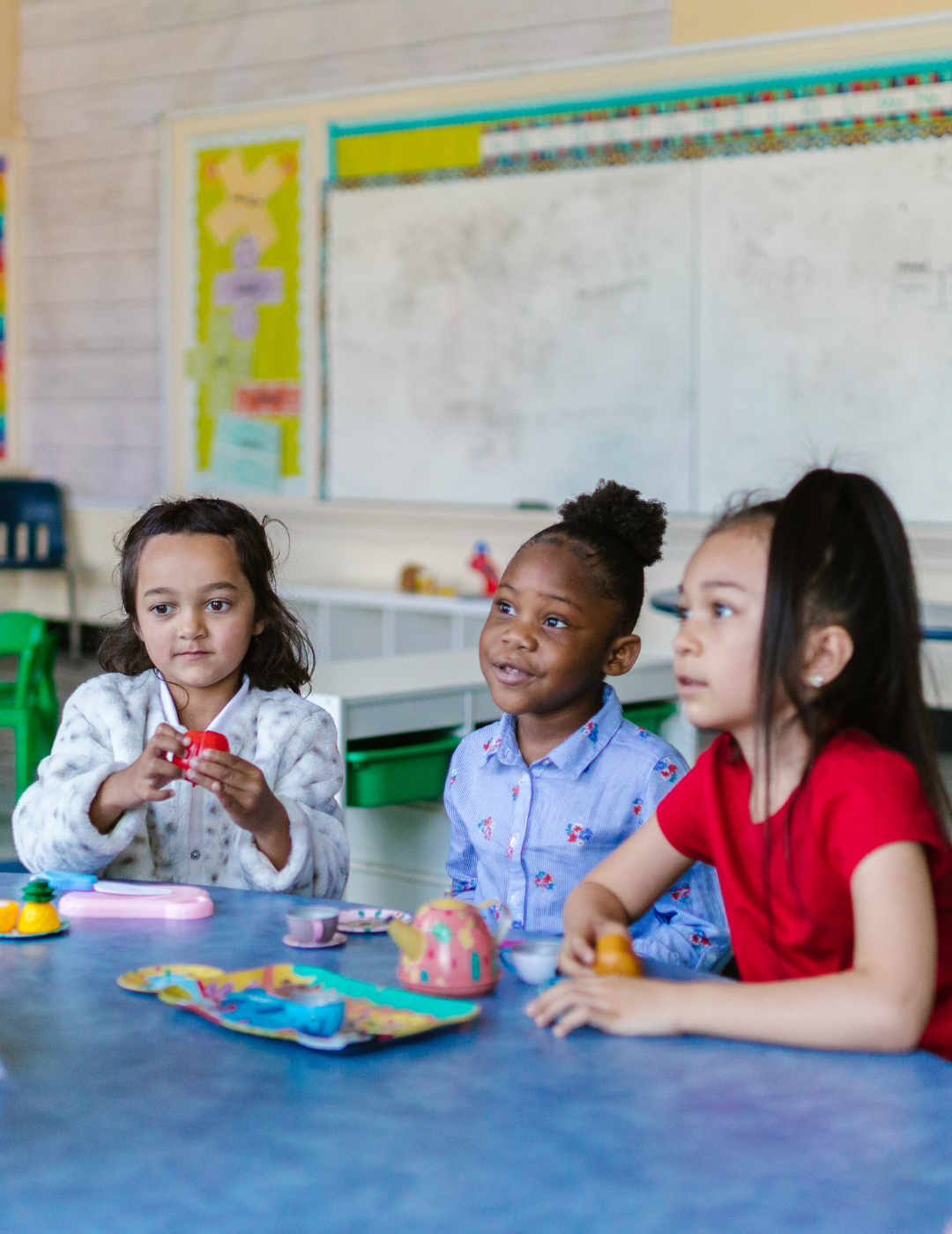 Three children sit at a school desk listening to a teacher.