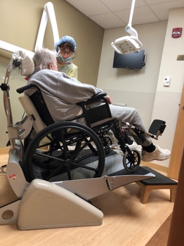 A patient sits in a wheelchair that is tilted backward on a small tilted platform, with their head supported by a headrest attached to the device.