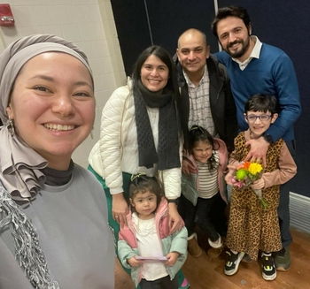 A woman smiles near the camera. Behind her, two men and a woman lean together side-by-side. Three small children stand in front of them.