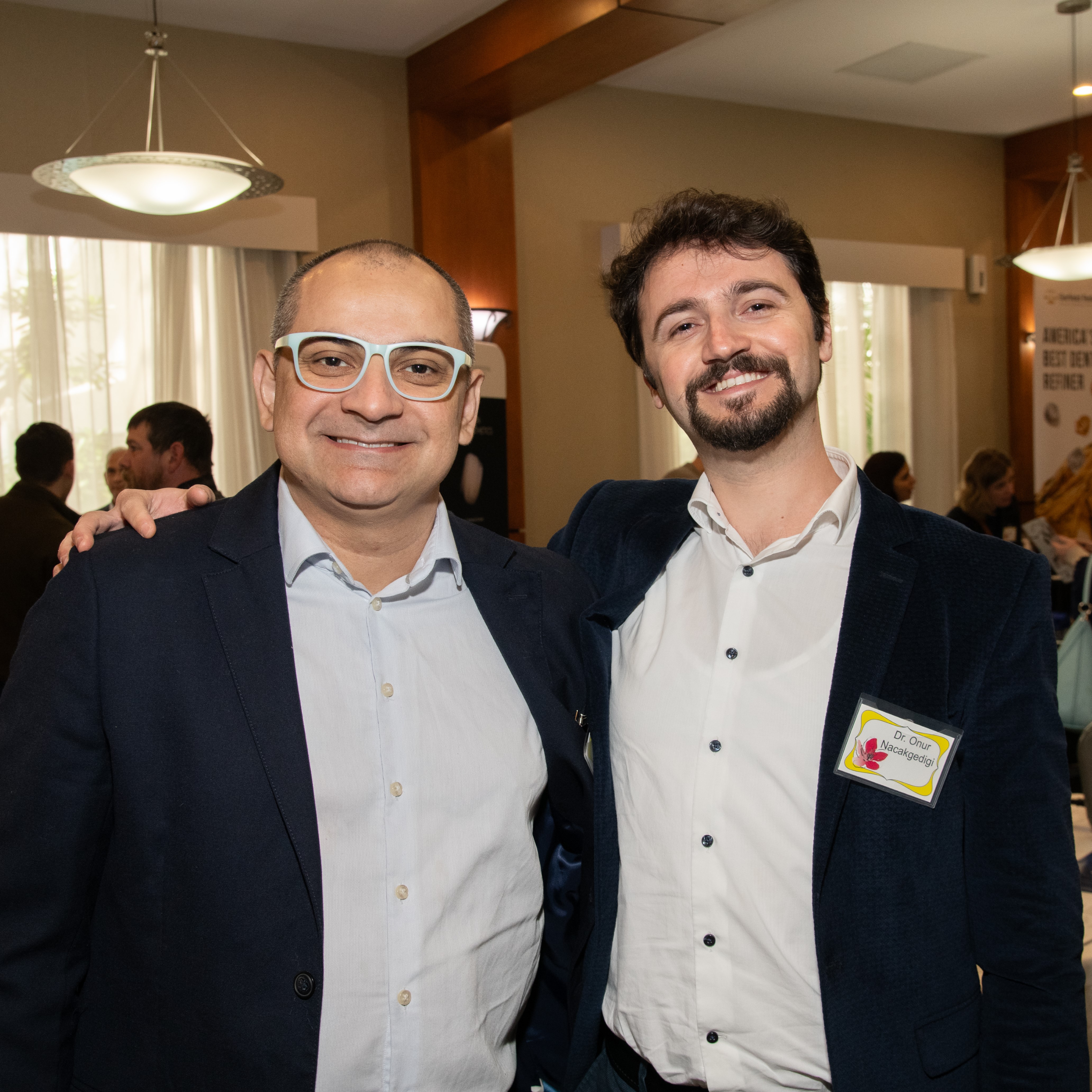 Two men in suits smile stand side-by-side. One has his arm around the other's shoulder.
