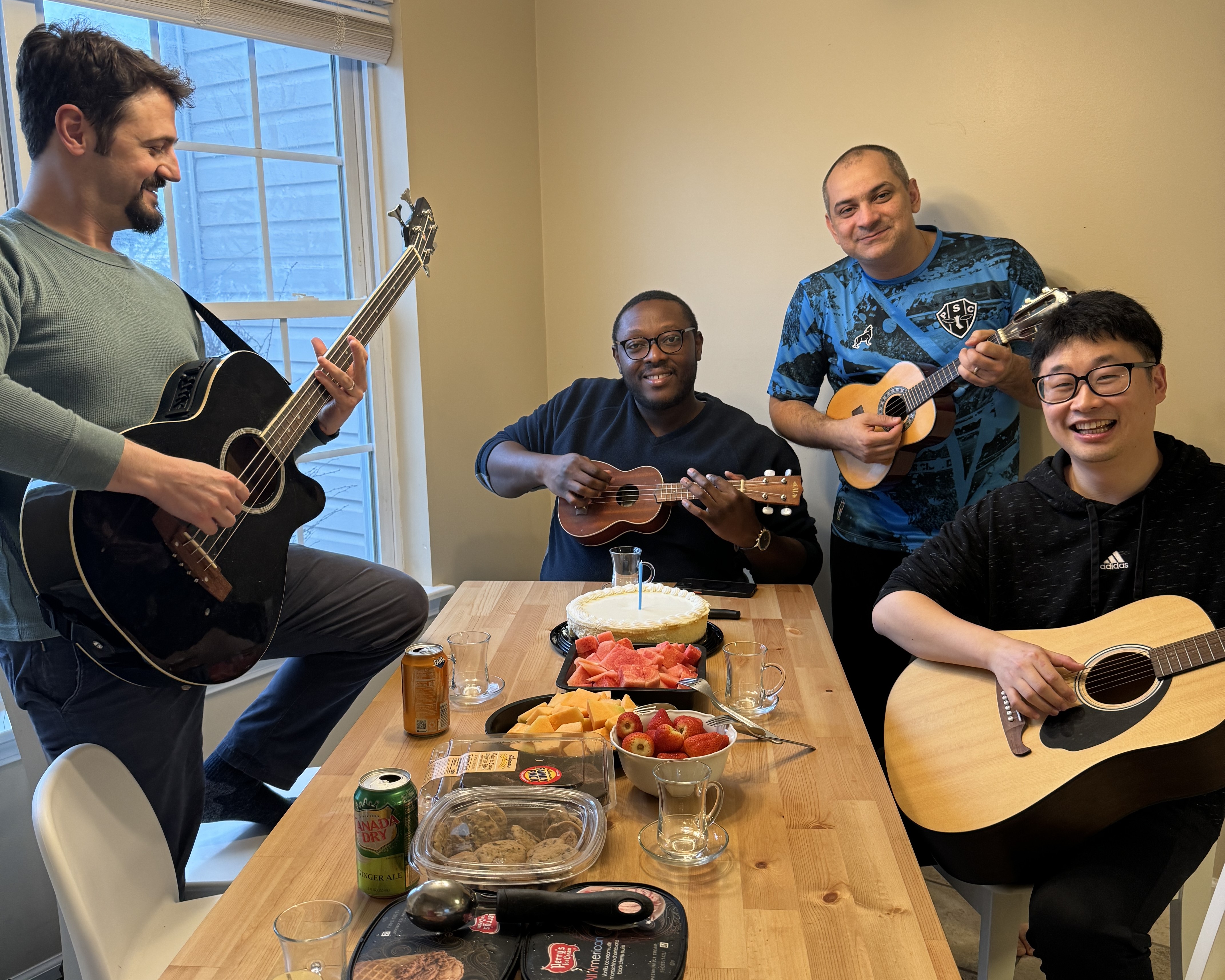 Four people gather around a table with platters of food on top of it. Two are playing ukuleles and the other two are playing acoustic guitars. 