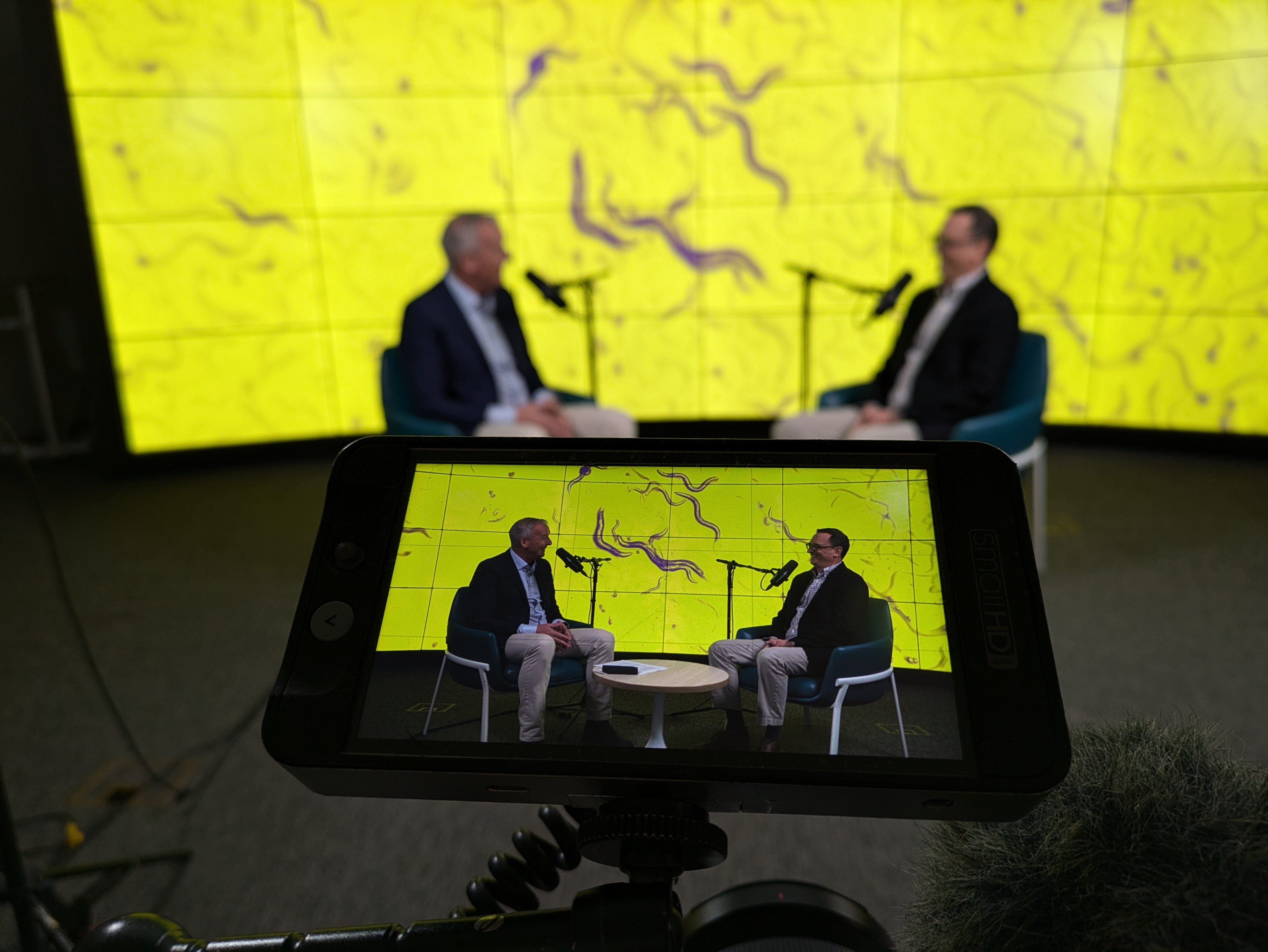 Two people sitting in chairs talking with microphones and cameras facing them.