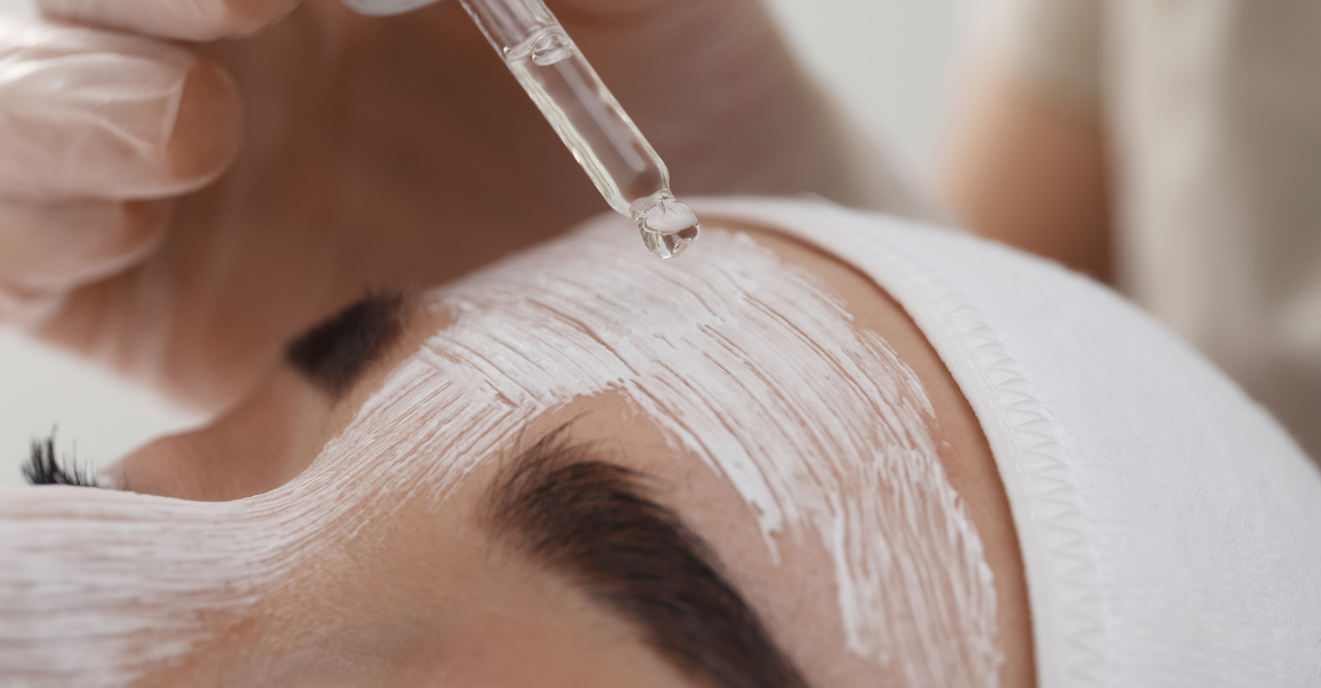 Close up of forehead with white cream on it. Hand uses dripper to add clear liquid product to forehead. 