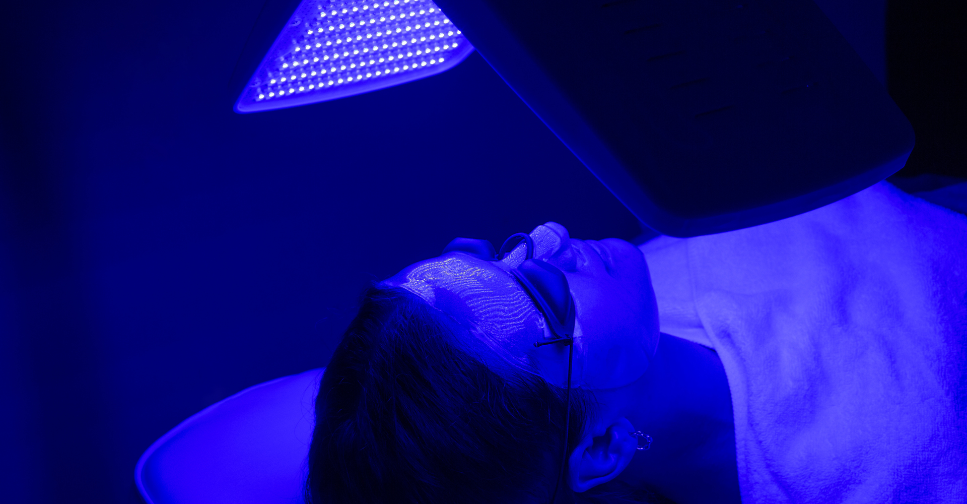 Woman lays in dark room with facial mask on. A blue LED light shines on her face as a skin treatment.