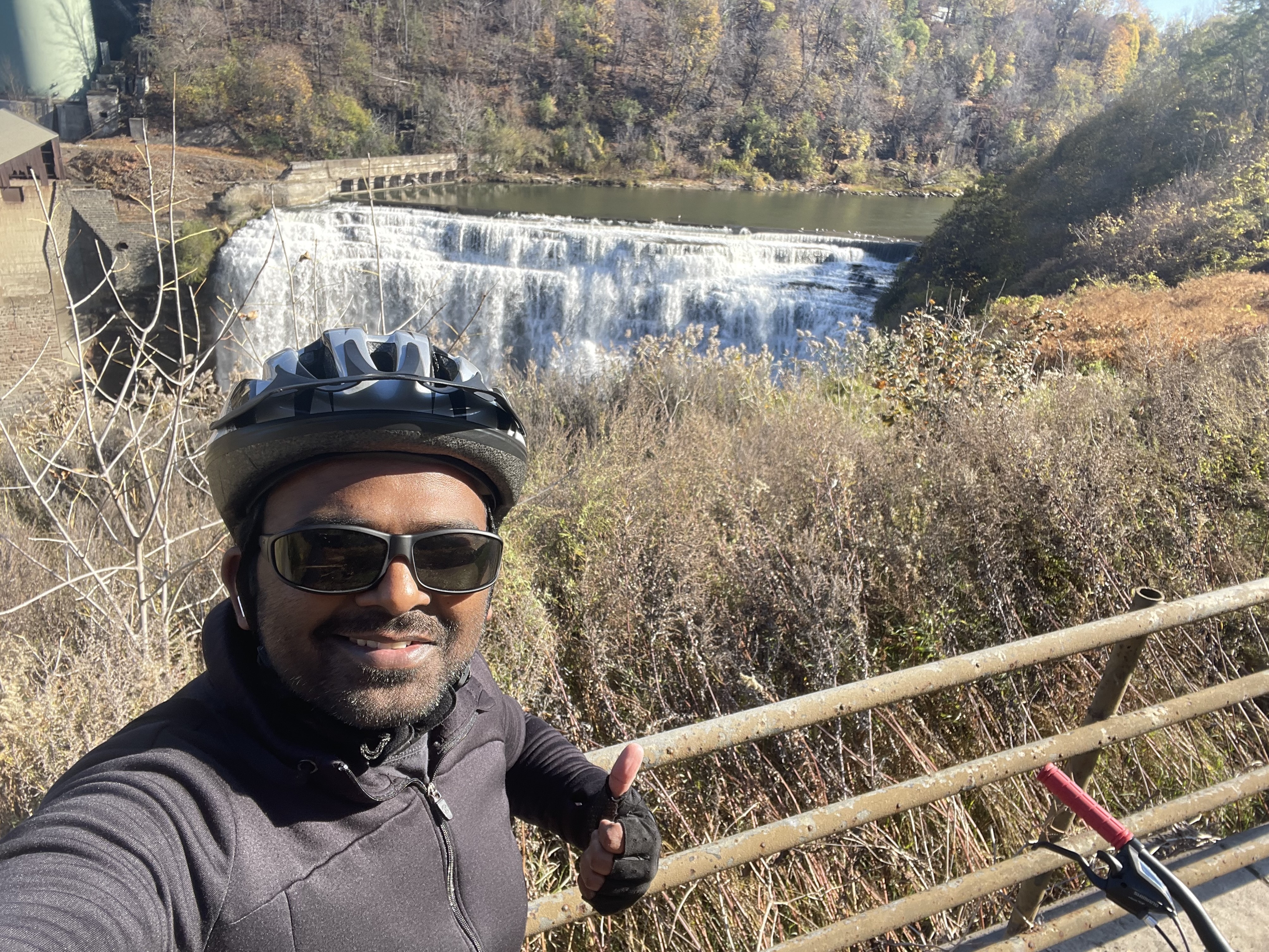 A man in a biking helmet and sunglasses gives a thumbs-up in front of a waterfall surrounded by autumn trees and plants.