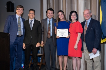 6 people stand in a row in professional attire. A woman near the middle holds a certificate.