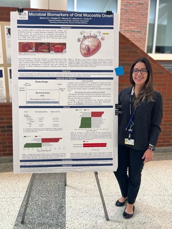 A woman stands next to a scientific poster titled "Microbial Biomarkers of Oral Mucositis Onset"