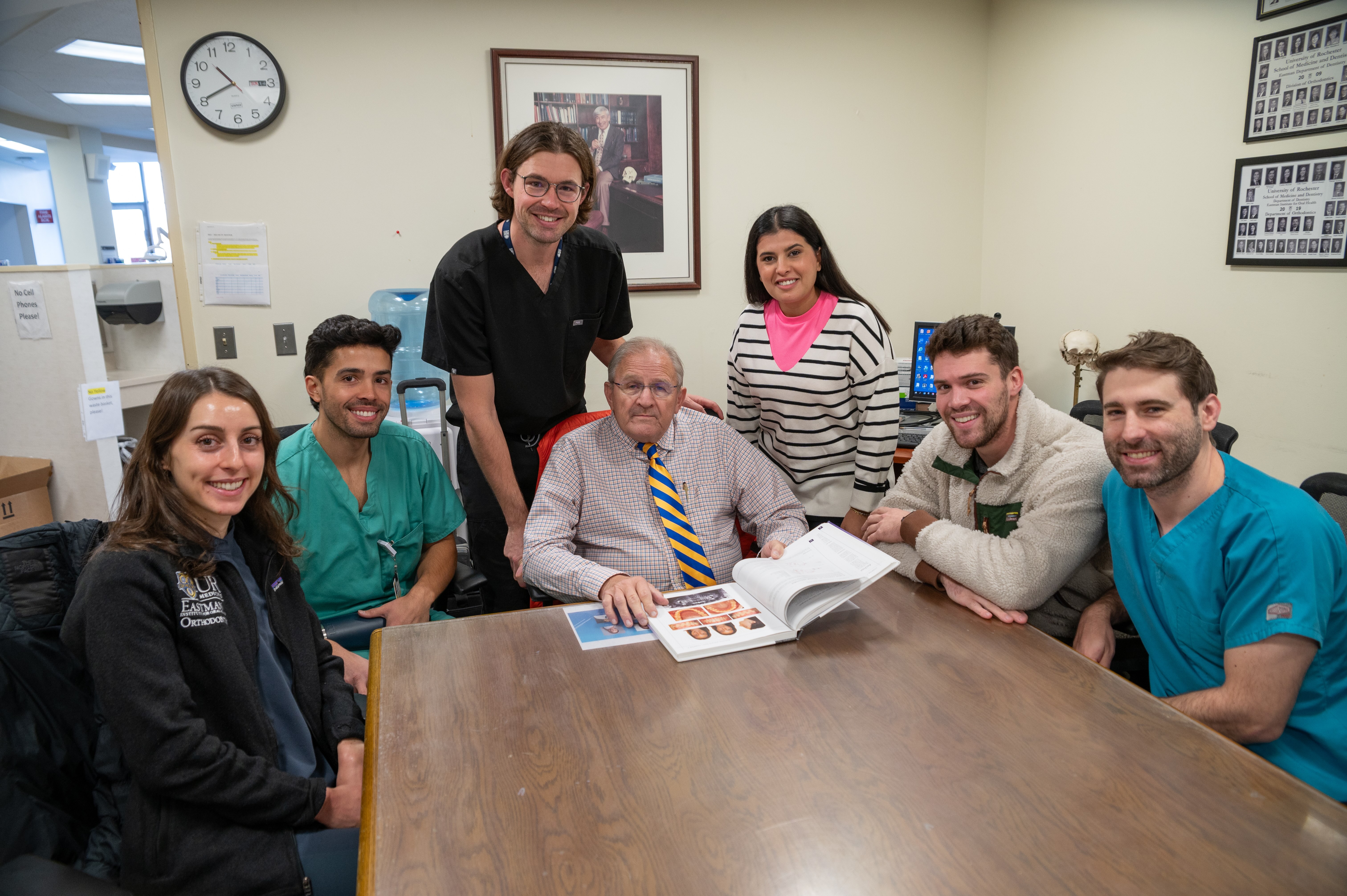 Six people, most of them wearing scrubs, surround an older man in a tie who is sitting at a table with an open textbook in front of him.