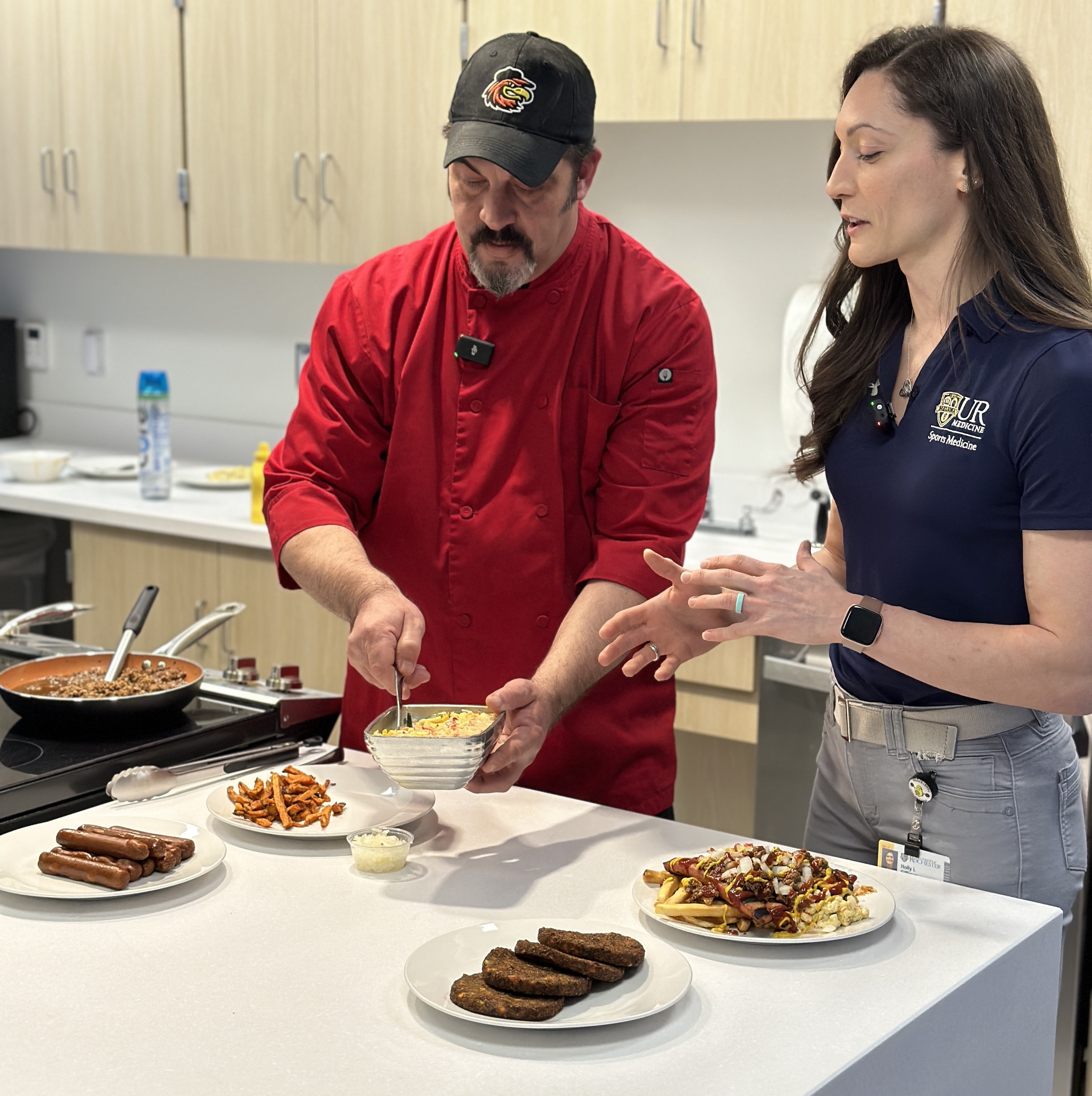 Woman and man stand in UR Medicine Fitness Science kitchen holding health garbage plate ingredients, including vegetable salad, sweet potato fries, bean burgers.