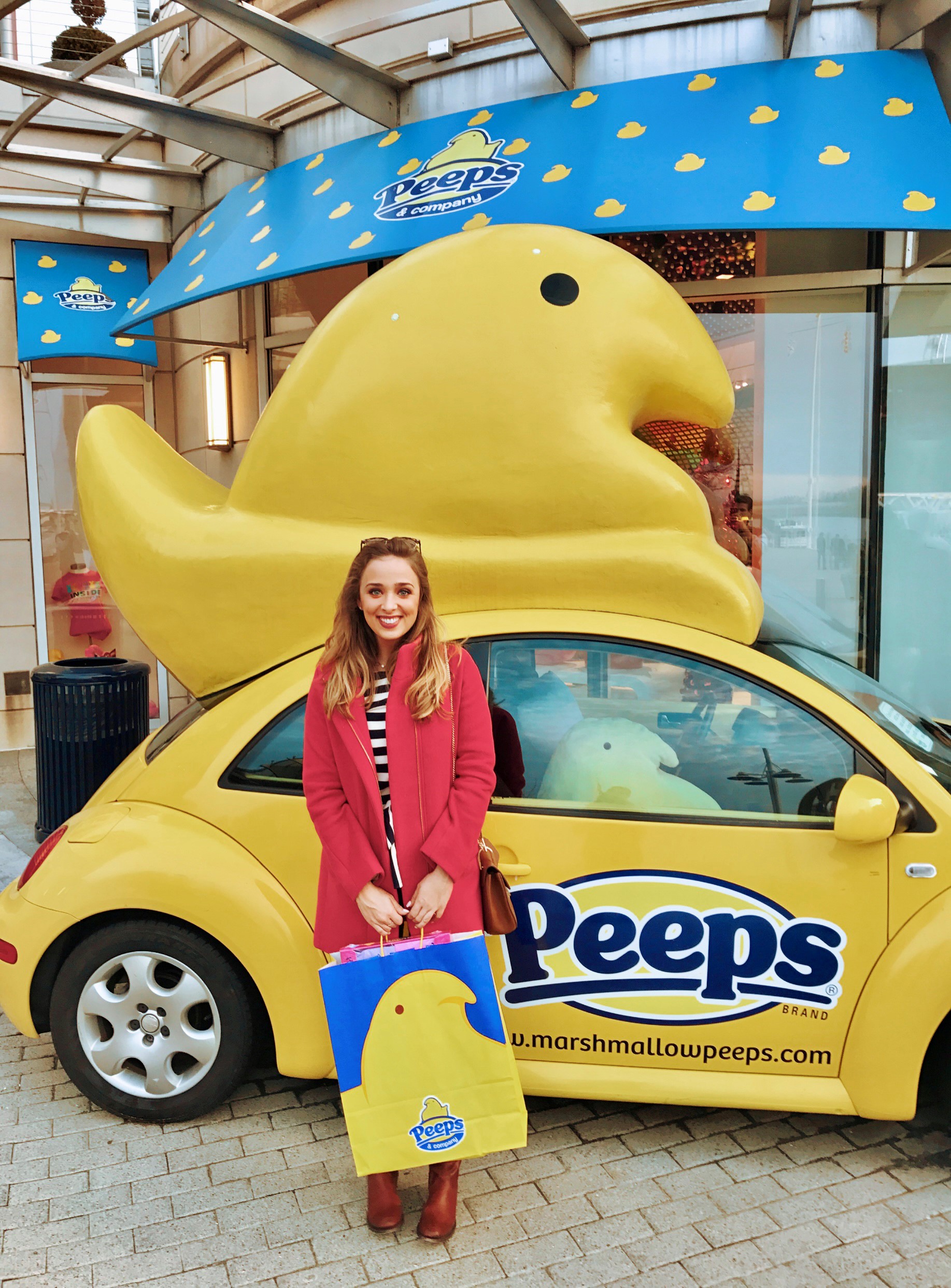 A smiling woman holds a large Peeps bag filled with treats, standing in front of a yellow car with a huge model of a marshmallow chick built onto the top of it and the Peeps logo on the side. There is a giant stuffed marshmallow peep sitting in the front seat of the car.