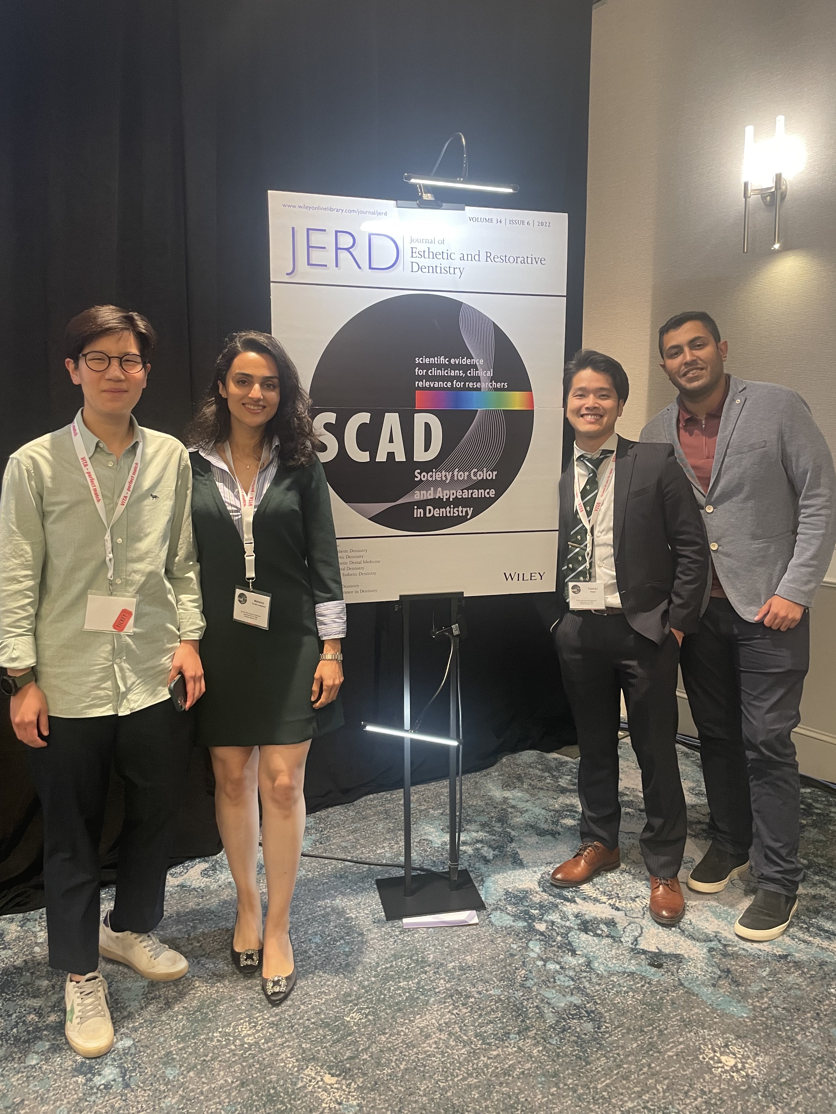 Four people in formal attire stand in front of a large sign that says "SCAD: Society for Color and Appearance in Dentistry"