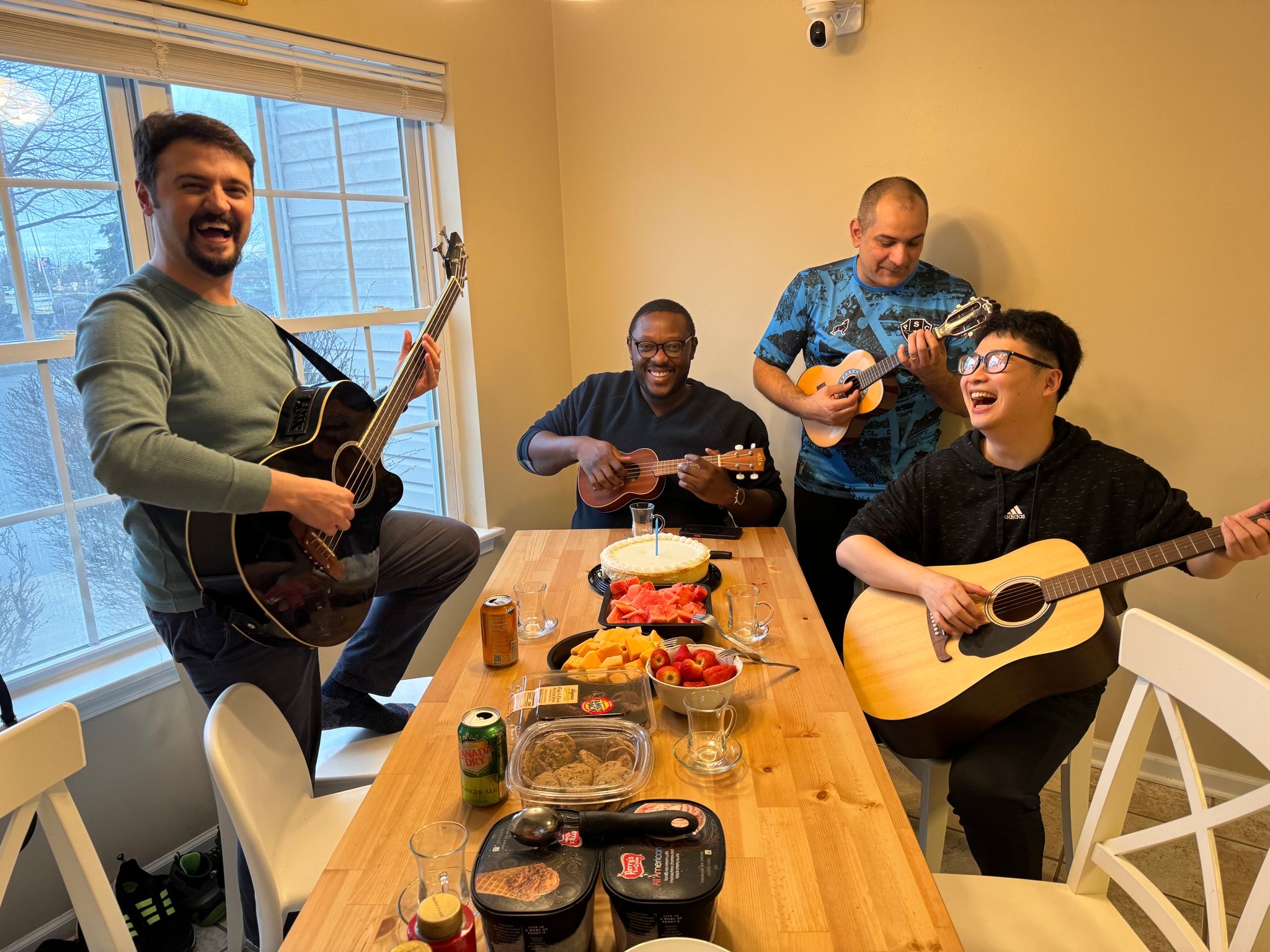 Four men sit laughing around a table that has fruit and desserts on it. Two men are playing guitars and the other two are playing ukuleles.