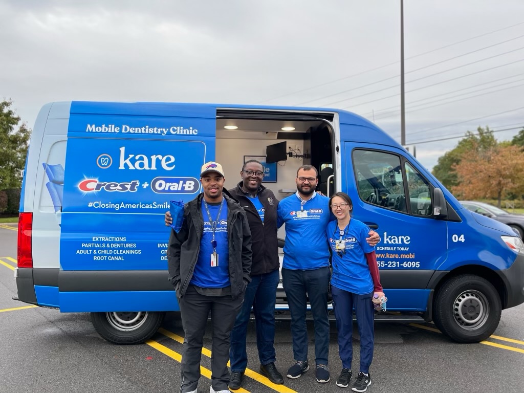 Four people wearing blue "Crest + Oral-B" t-shirts stand in front of a blue van labeled "Mobile Dentistry Clinic"