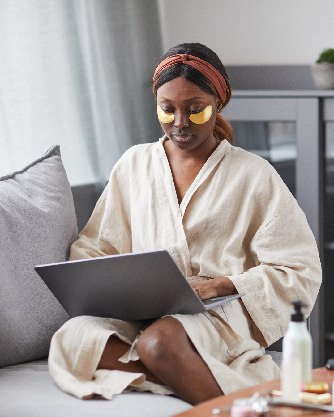 Woman sits on couch wearing eye patches underneath her eyes for her nighttime skin routine.