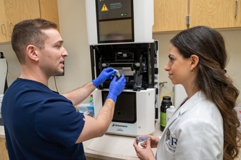 A person in scrubs and a person in a lab coat remove items from a 3D printer