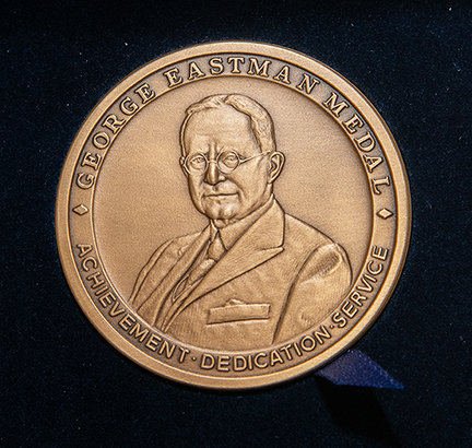 A close-up of a bronze-colored circular medal, with text reading "George Eastman Medal" and "Achievement, Dedication, Service" around the edge, and a portrait of George Eastman in the middle.