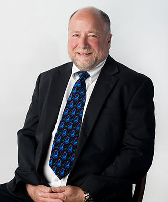 man sitting in a chair wearing a dark suit and a blue tie. hands are folded in lap.