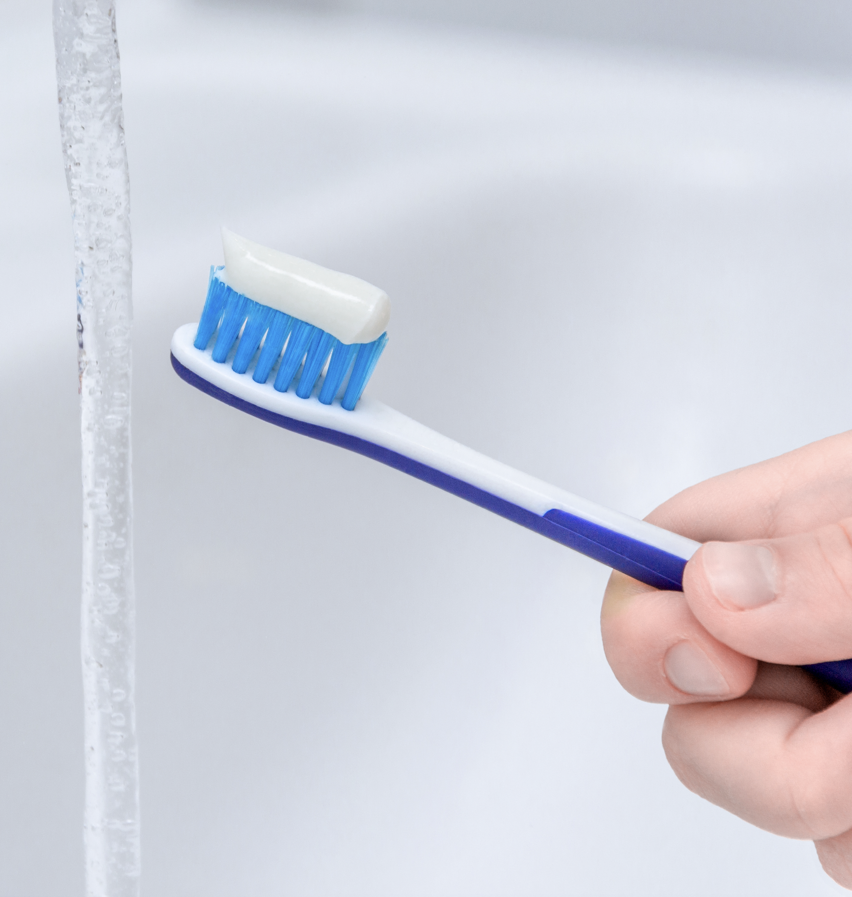 Hand holding toothbrush with toothpaste near running water