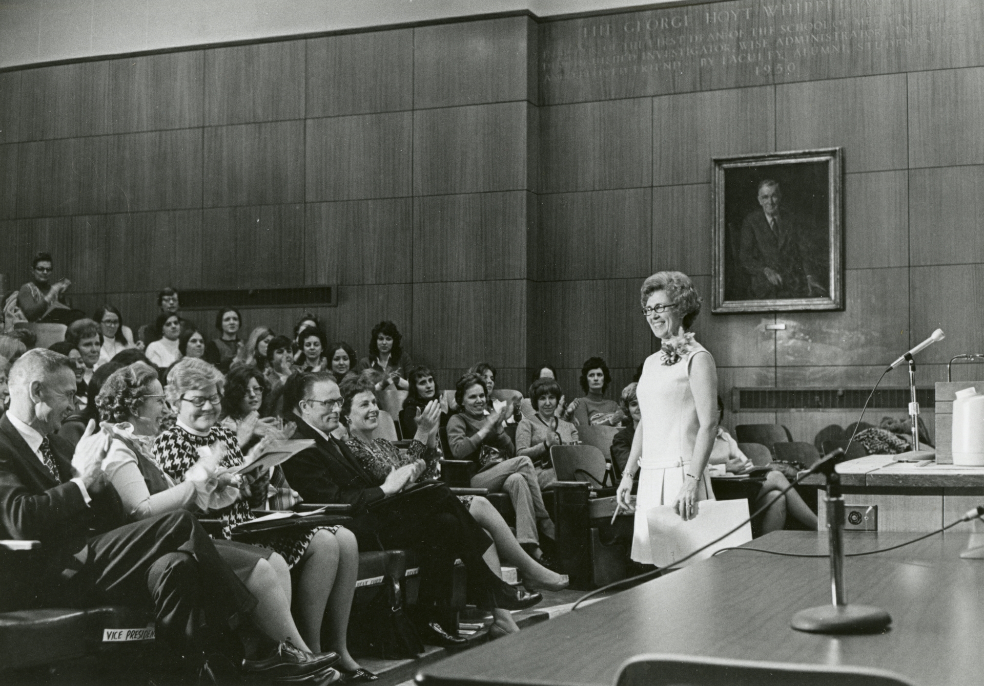 In a 1970s black and white photo, Loretta Ford smiles at the front of an auditorium while an audience cheers for her.