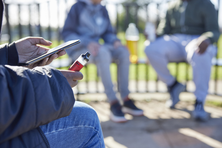 Teen holding cell phone and e-cigarette in a park.