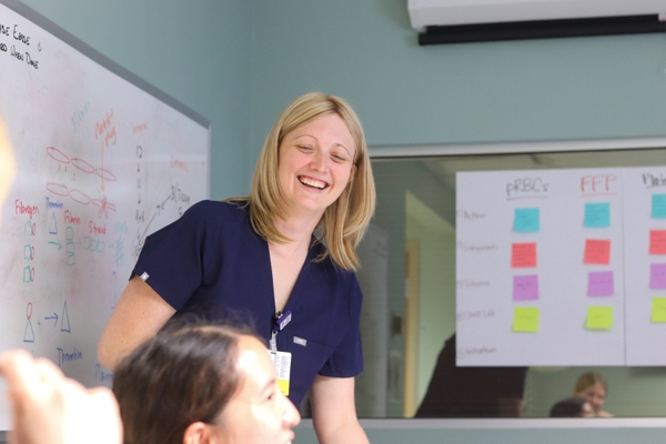 Dr. Courtney Olson-Chen smiles and looks at learner while standing in front of a white board and a large poster with colorful post-it notes