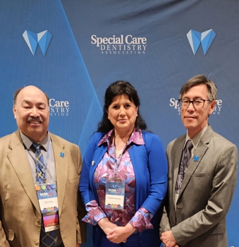 A woman in a blue cardigan stands between two men in suits. Behind them is a backdrop that reads "Special Care Dentistry Association".