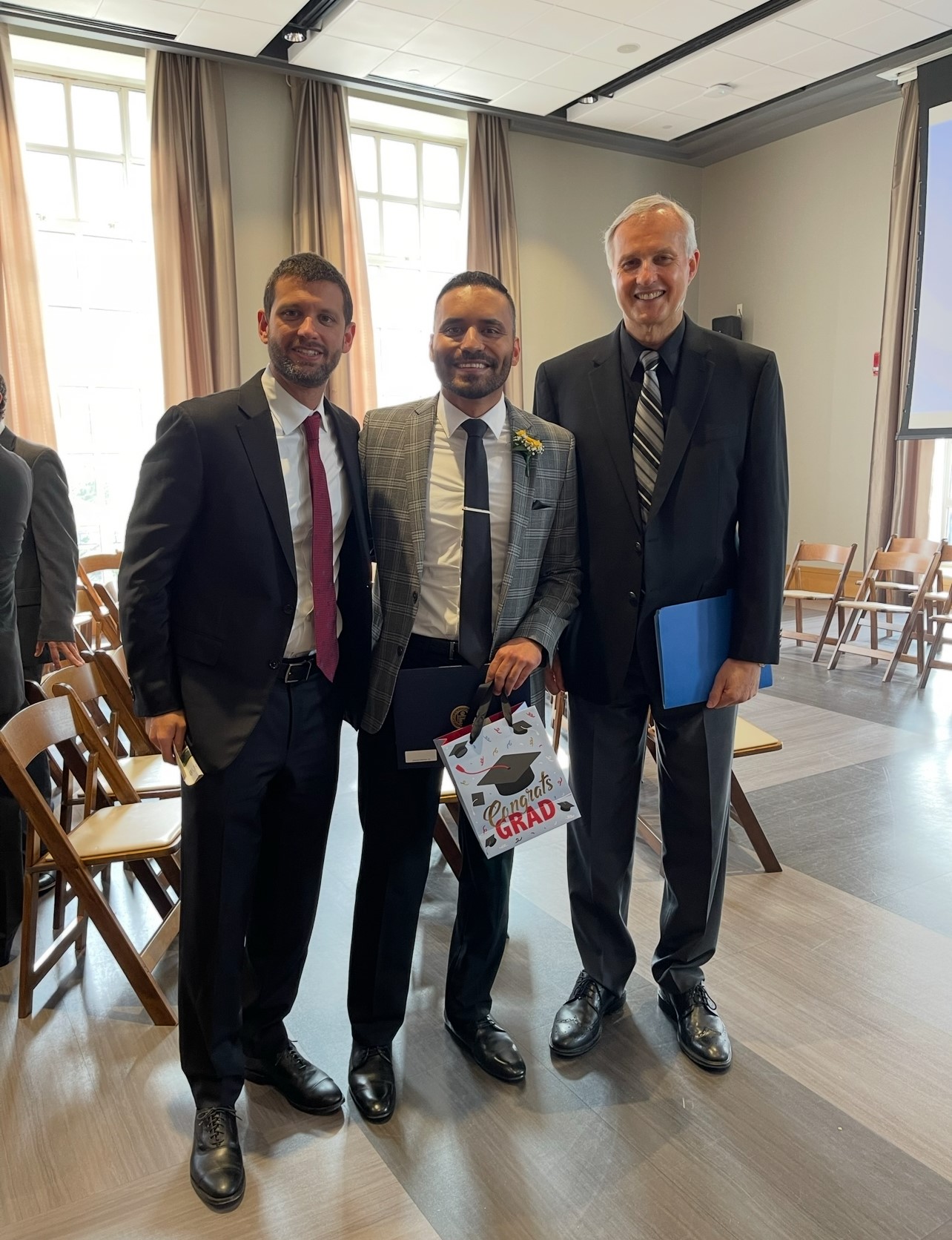 Three men in suits and ties. The one in the middle holds a diploma and a bag that says "Congrats grad".