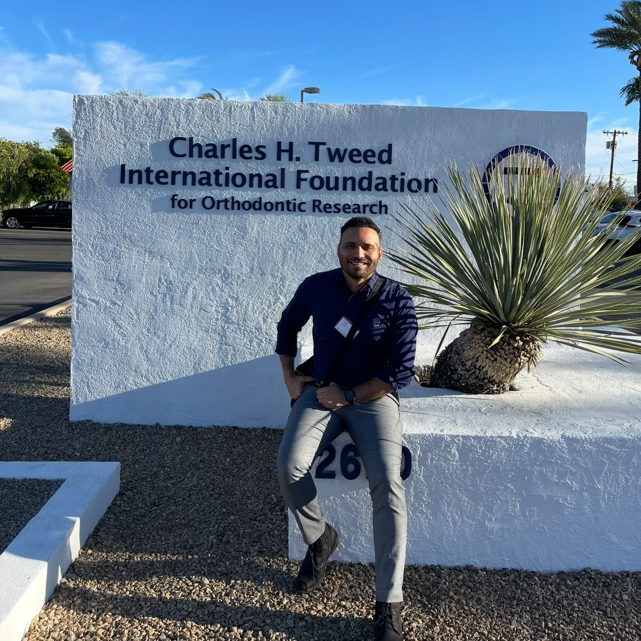 A man sits in front of a concrete sign reading "Charles H. Tweed International Foundation for Orthodontic Research".