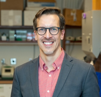Smiling individual in glasses and a blazer standing in a lab.