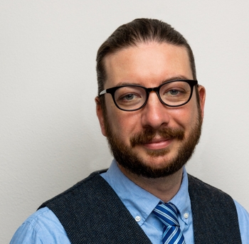Individual with glasses wearing a tie and vest, smiling in front of a plain background.