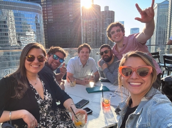 Two women in sunglasses sit in front of a table, with 4 men sitting behind them waving and smiling.