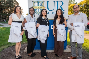 5 people hold white lab coats and stand in front of a sign that reads "UR Medicine".