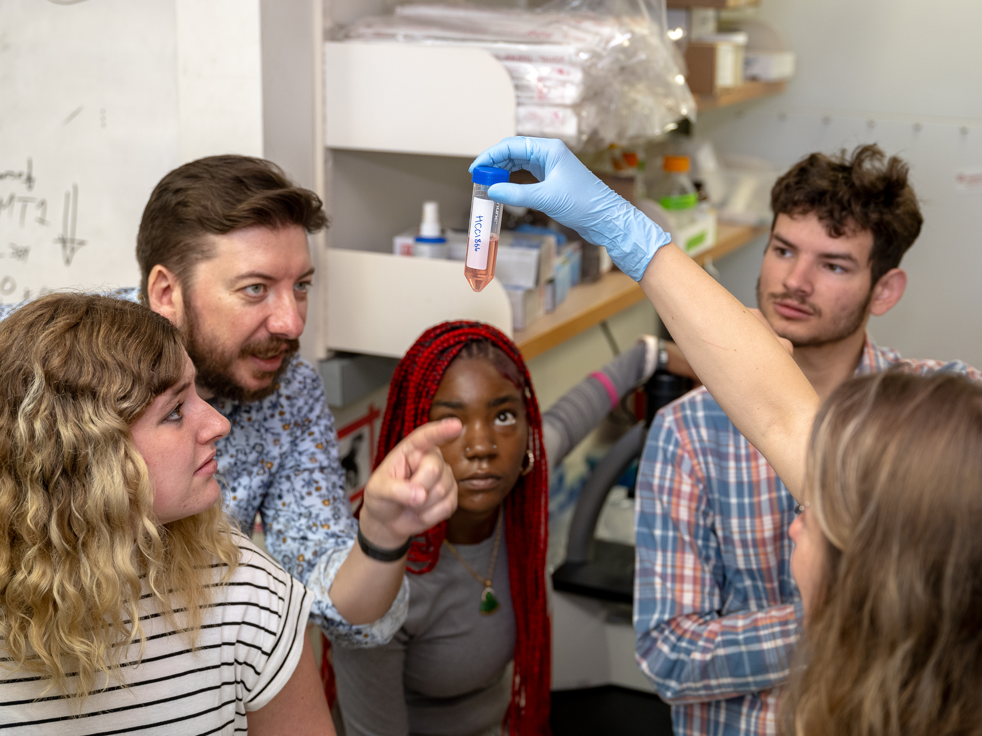 Wyatte Hall, PhD, points to a clump of cells at the bottom of a centrifuge tube being held up by Sara Blick-Nitko, PhD, as three high school students look on.