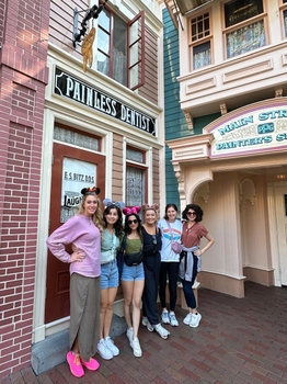 a group of women wearing Mickey Mouse ears pose in front of a storefront with a sign reading "Painless Dentist".