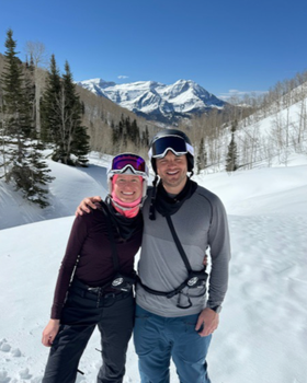 A man and woman in skiing goggles stand in front of a snowy mountain.