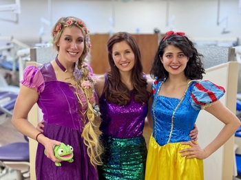 3 women stand together in costumes in a dentistry clinic. They are dressed as Rapunzel, Ariel, and Snow White.