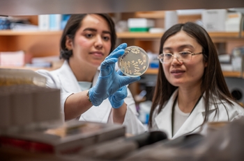 Two women in lab coats hold up a petri dish of bacteria to examine.