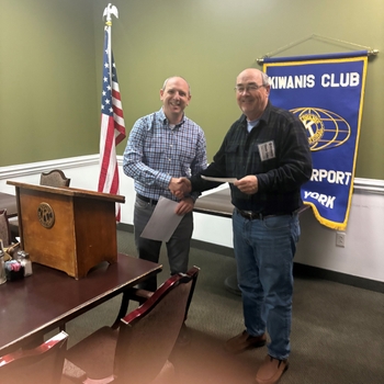 Two men shake hands in front of a banner that reads "Kiwanis Club".