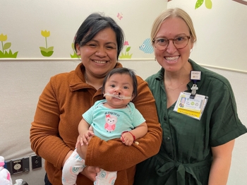 Dr. Shope poses next to a mother holding a baby with cleft lip and palate. The baby has a NAM device fit around their cleft which is held in place with medical tape.